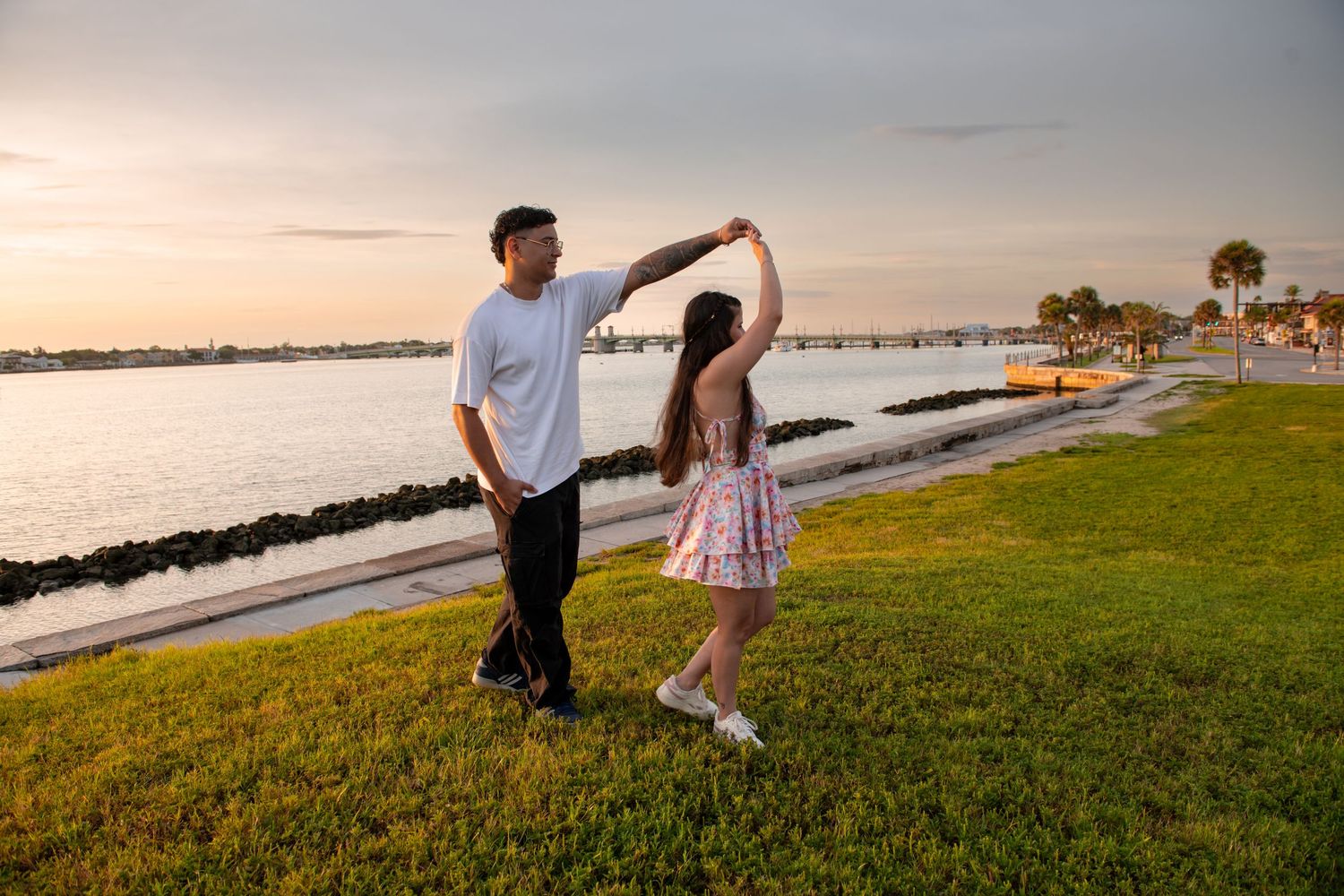 Couple dancing at sunset by the waterfront on green grass, golden hour light reflecting on calm bay waters.