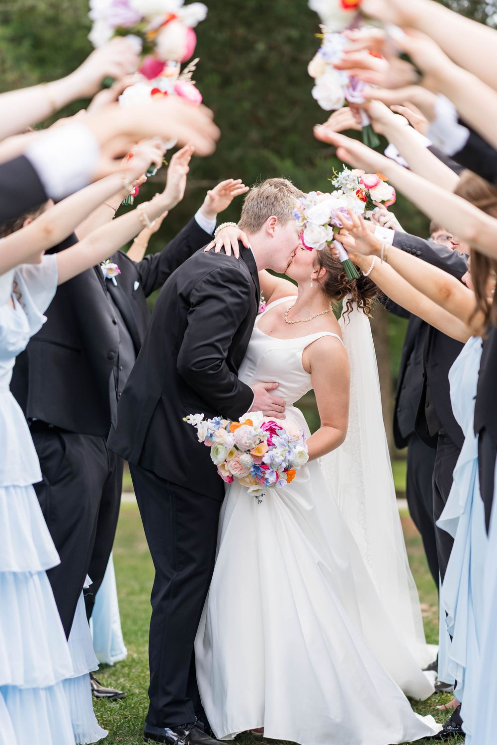 Bride and groom kissing under an arch of raised bouquets held by bridesmaids in blue dresses and groomsmen in black suits.