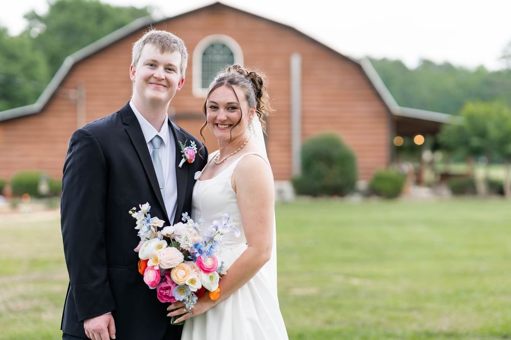 Smiling bride and groom pose outdoors in front of a rustic barn venue, bride holding a colorful bouquet.