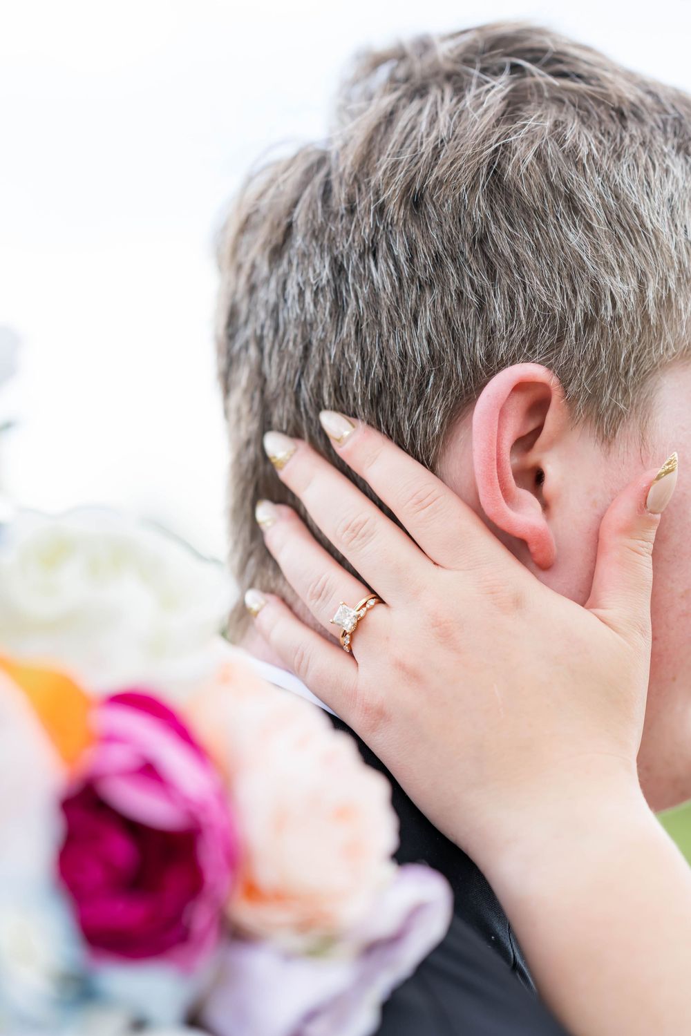 Bride wearing a gold diamond engagement ring gently touching grooms face near colorful wedding bouquet.