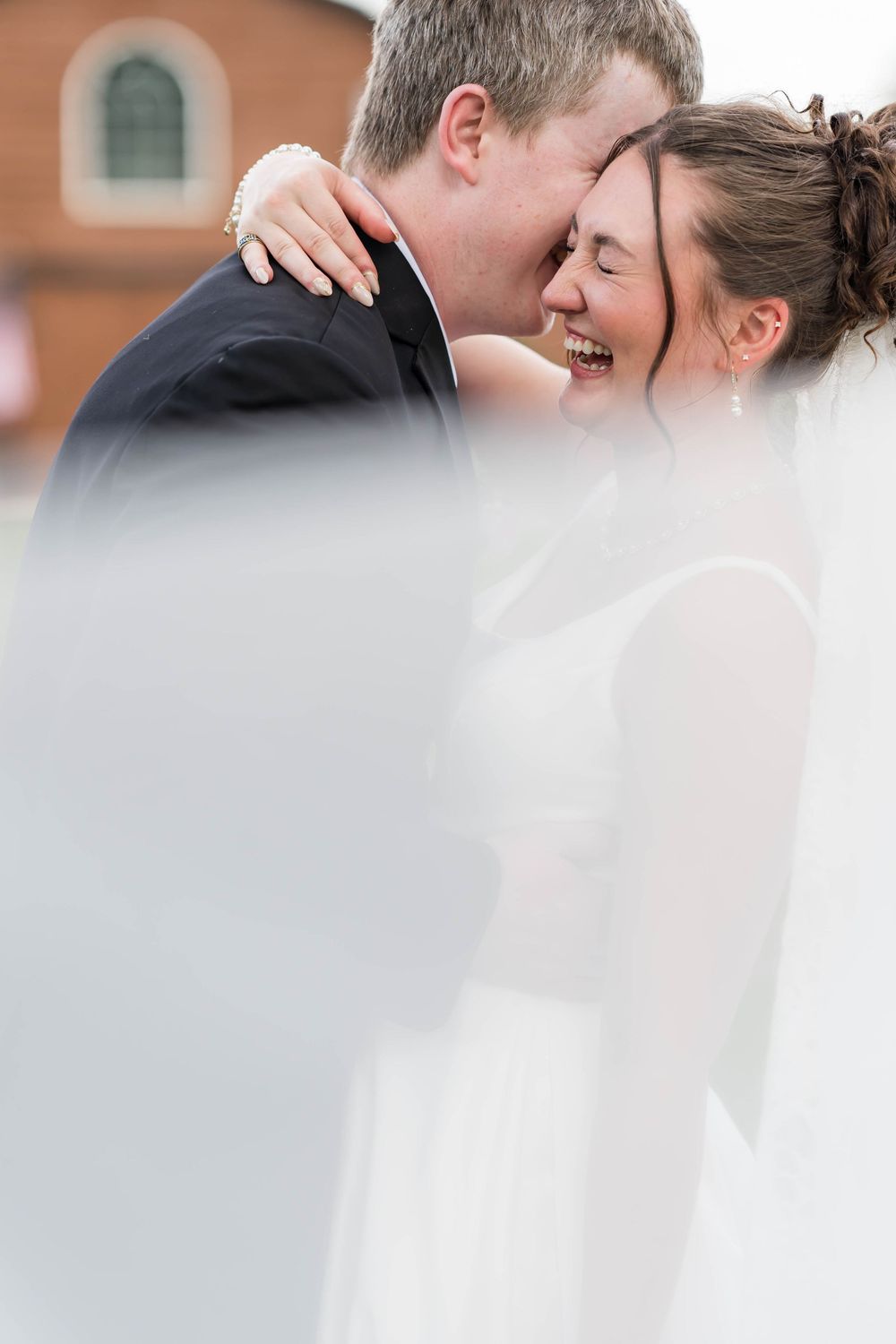 Bride and groom sharing a joyful moment under a wedding veil, laughing and embracing outdoors.