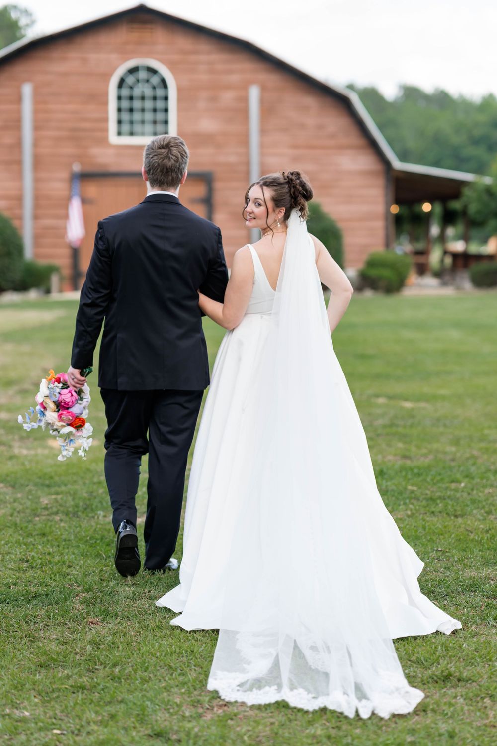 Bride in white gown and groom in black tuxedo walking toward rustic barn venue, bride glancing back over shoulder.