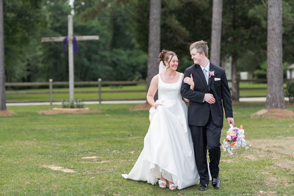 Bride and groom walking together on green lawn near trees with cross in the backgrou, smiling, bride in white gown holding colorful bouquet.