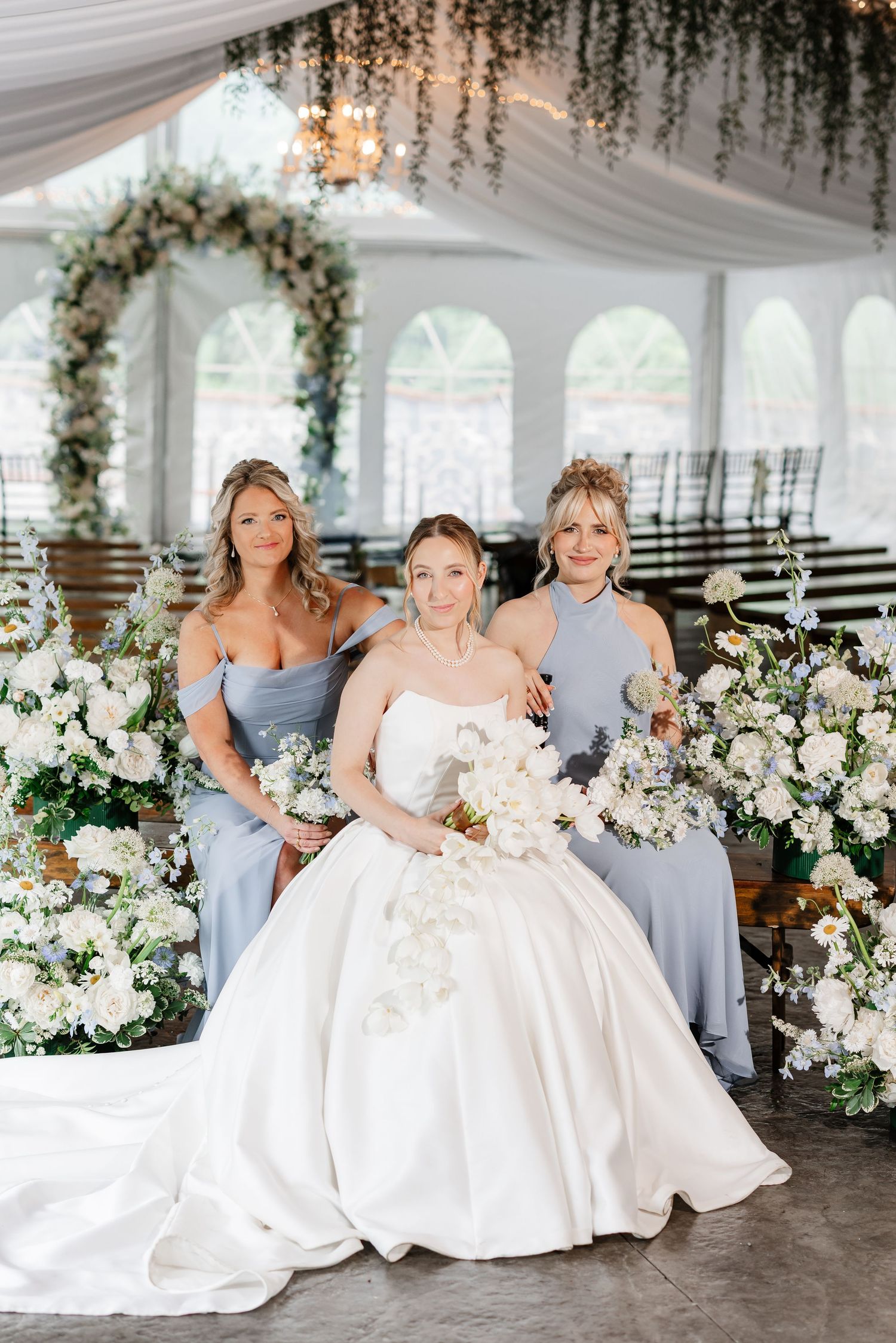 Bride in white ball gown seated between two bridesmaids in dusty blue dresses inside floral decorated wedding tent.
