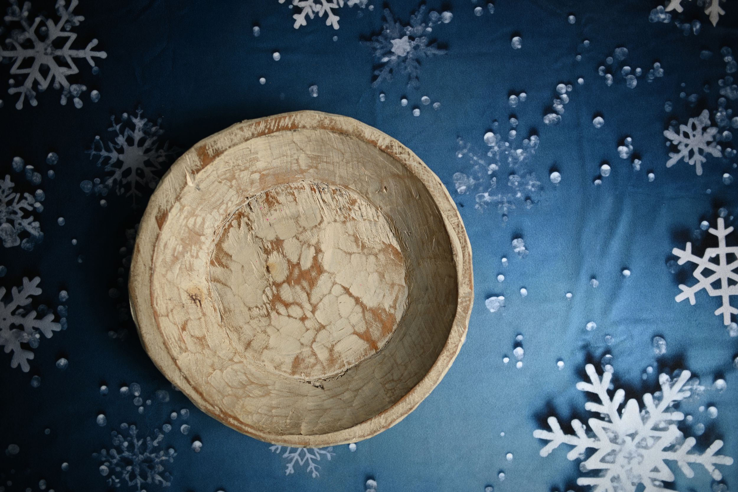 Rustic wooden bowl with cracked texture on blue fabric surrounded by white decorative snowflakes.