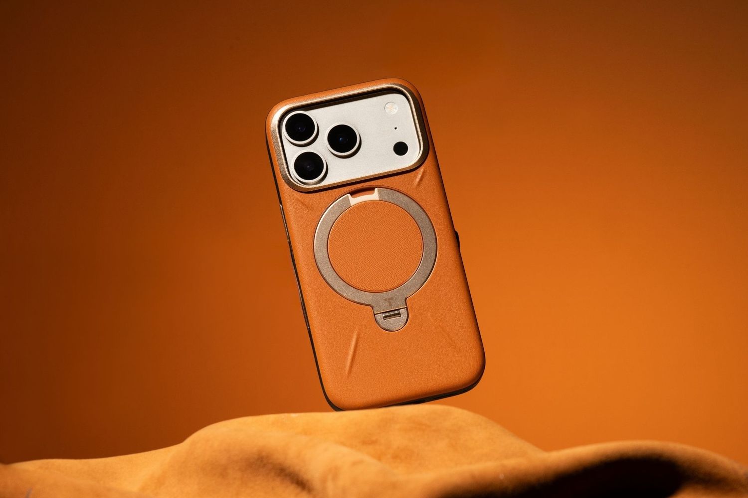 Orange leather MagSafe iPhone case with ring holder floating against warm amber background.