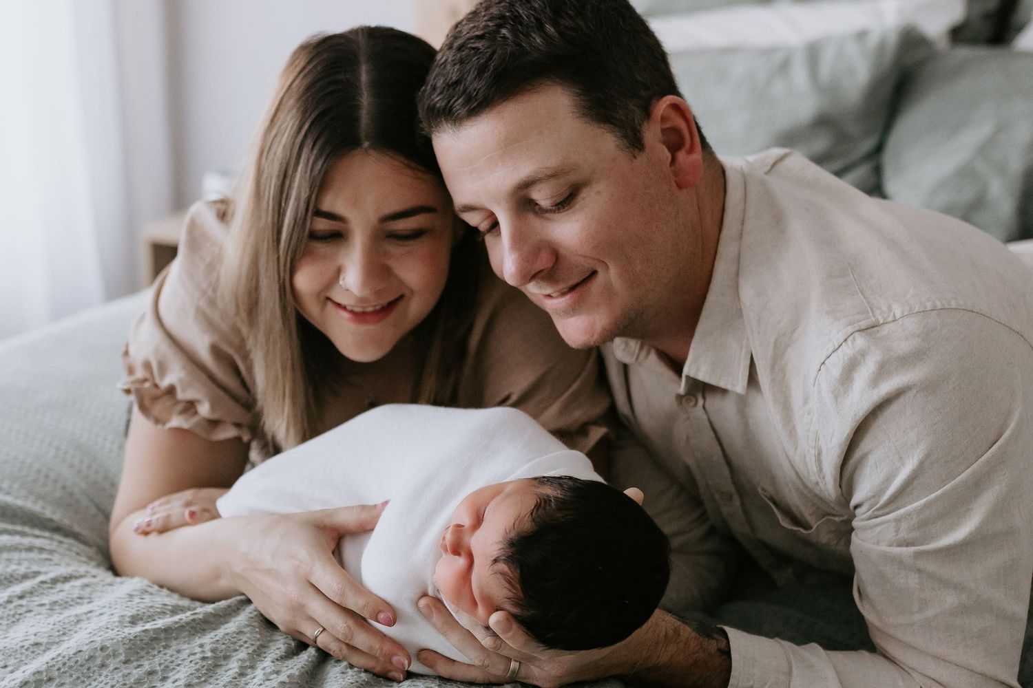 New parents smiling lovingly at their swaddled newborn baby while lying on a bed together.