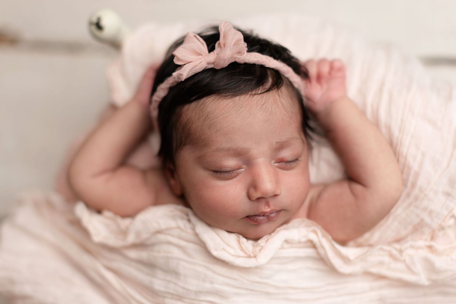 Peaceful newborn baby girl sleeping in pink wrap with bow headband in soft studio portrait photography.