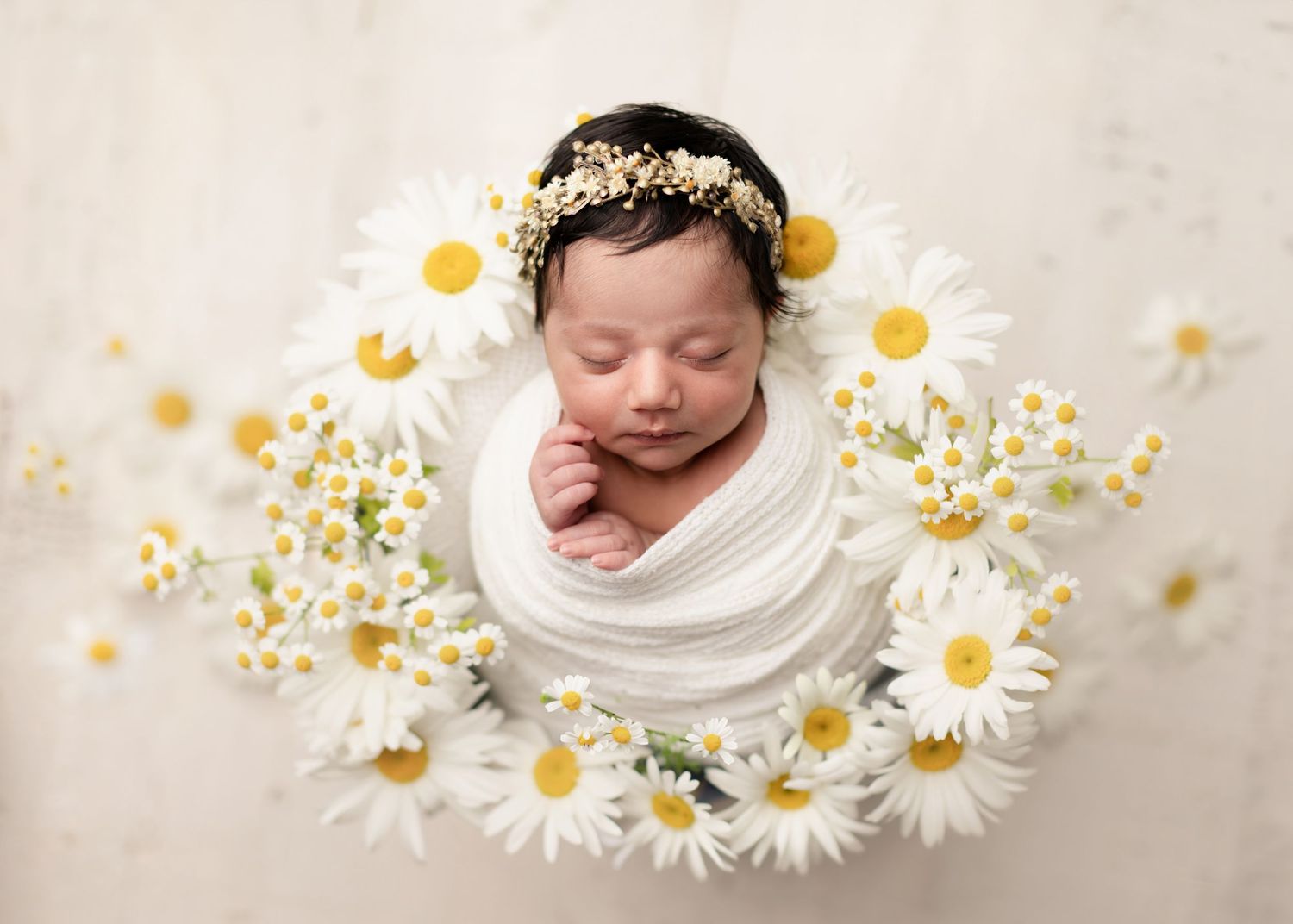 Newborn baby wrapped in white swaddled in a bed of daisy flowers wearing a floral headband.