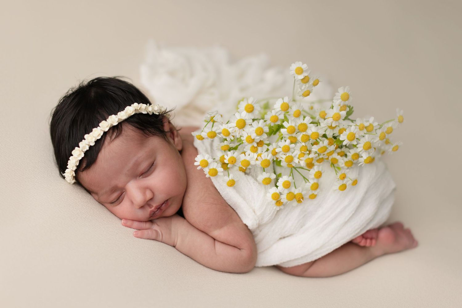 Sleeping newborn baby wrapped in white cloth with daisy flowers, wearing a pearl headband, on a beige background.