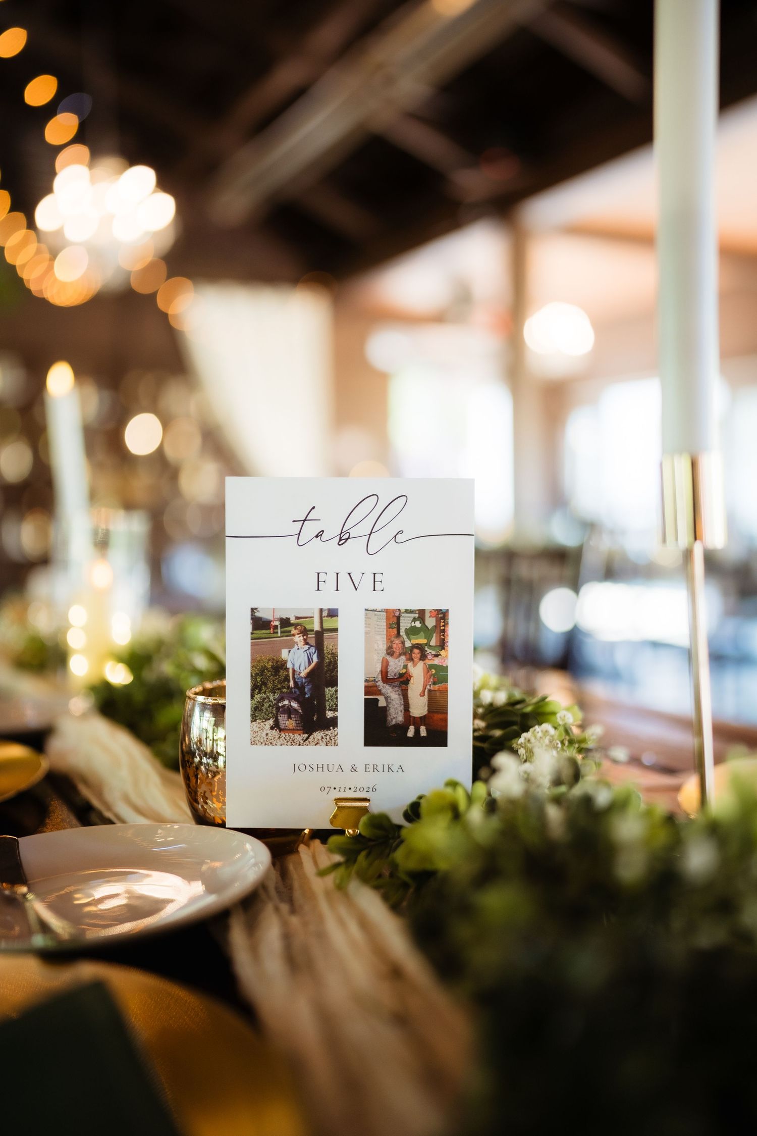 Elegant wedding table five card with couple photo, greenery centerpiece, and warm candlelight at reception.