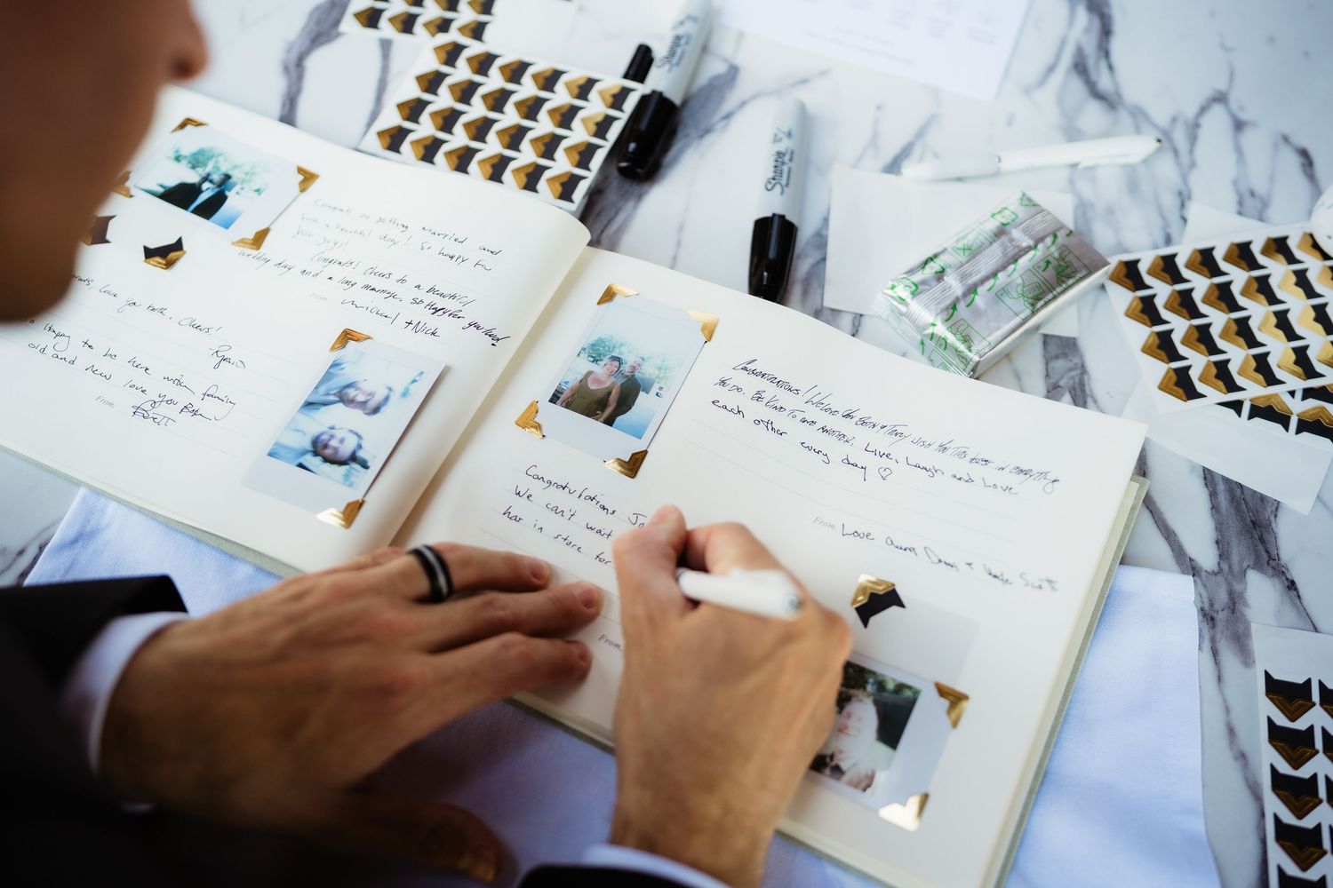 A person wearing a ring writes notes in a photo album scrapbook with polaroid pictures and pens on a marble surface.