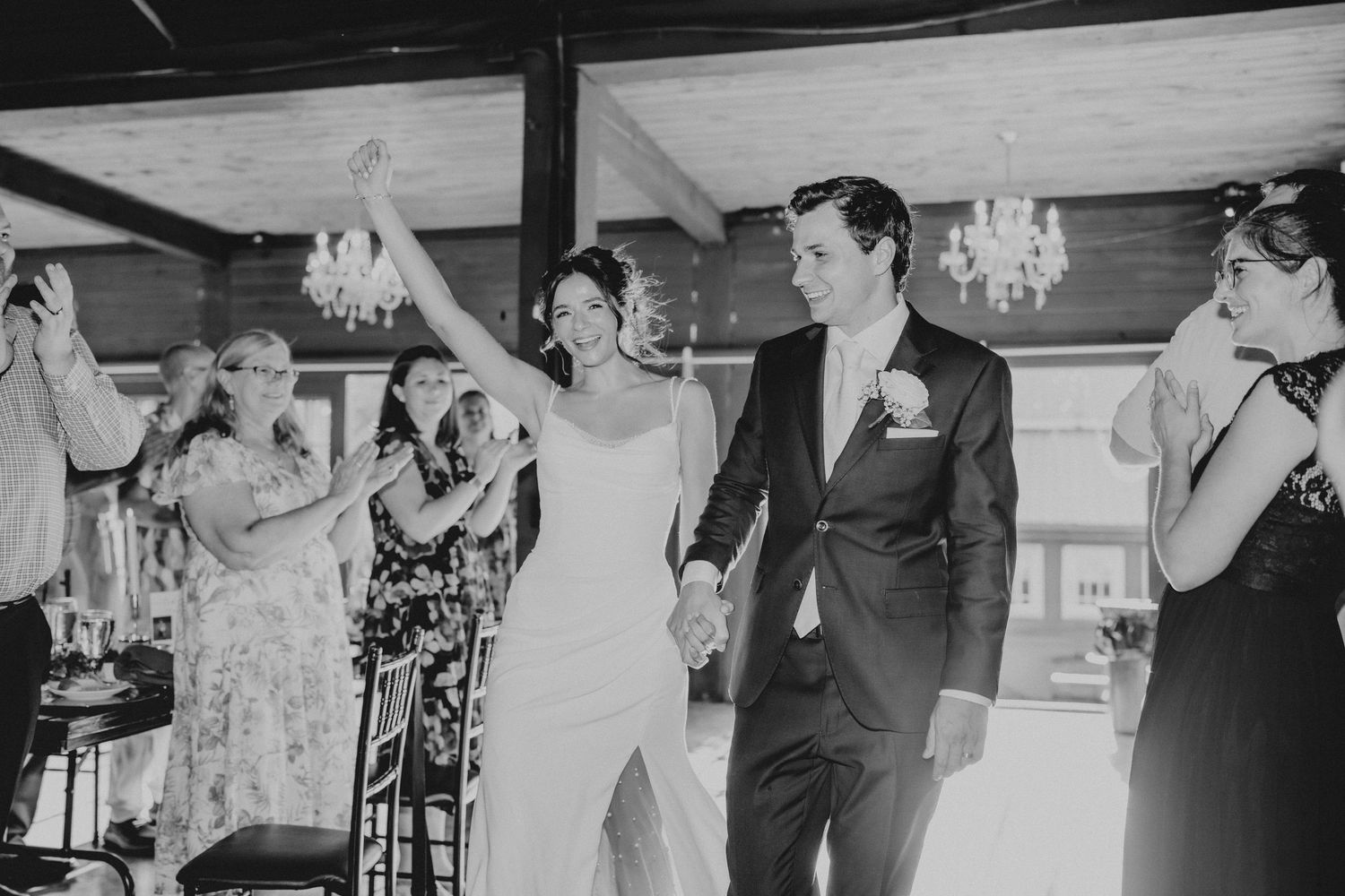 Bride and groom hold hands smiling as wedding guests celebrate around them in elegant black and white photo.