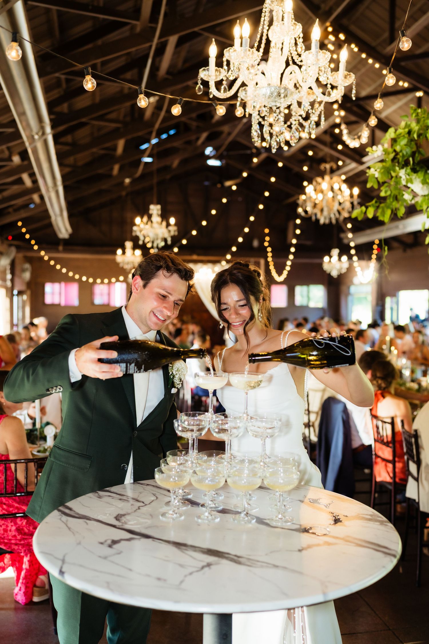 Bride and groom pouring champagne tower at wedding reception with crystal chandeliers and string lights.