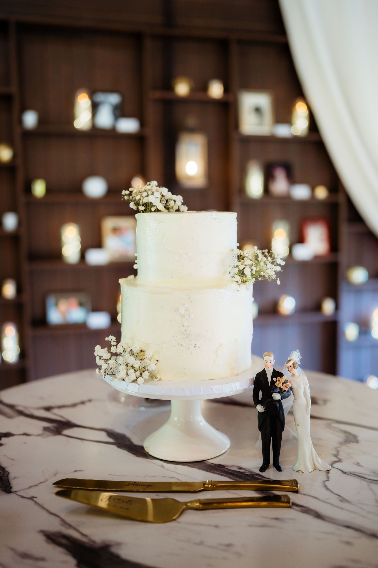 Two-tier white wedding cake with baby's breath on marble surface, bride and groom figurines, gold cake servers.