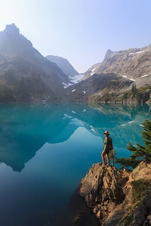 Hiker standing on rocky outcrop overlooking stunning turquoise alpine lake surrounded by rugged mountain peaks and glaciers.