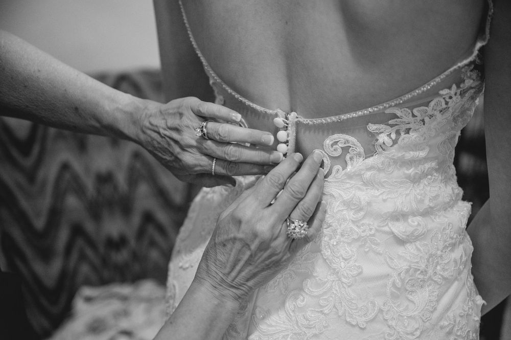 Hands fastening pearl buttons on the back of a lace wedding dress in black and white photo.
