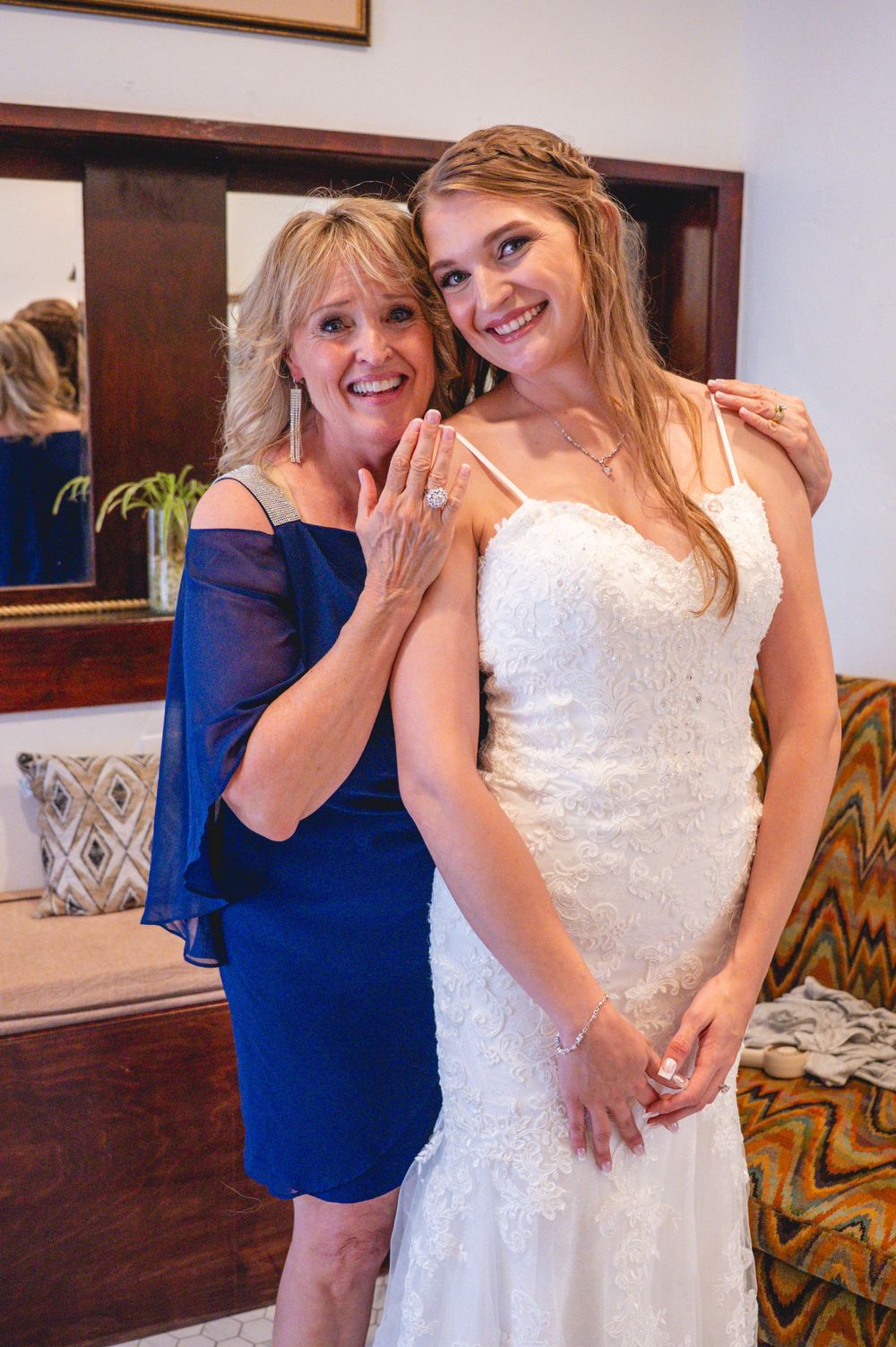 Bride in white lace gown poses with woman in blue dress showing off ring before wedding ceremony.