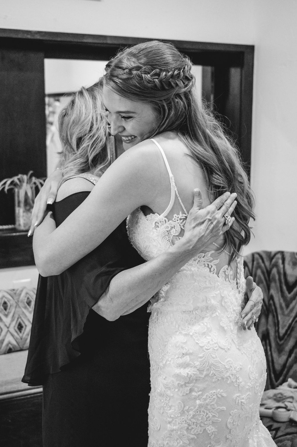 Black and white photo of a bride in lace gown with braided crown hugging a woman in black dress on wedding day.