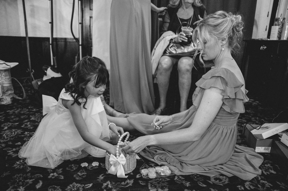 Bridesmaid helps flower girl with shoes at wedding preparation in black and white photo.