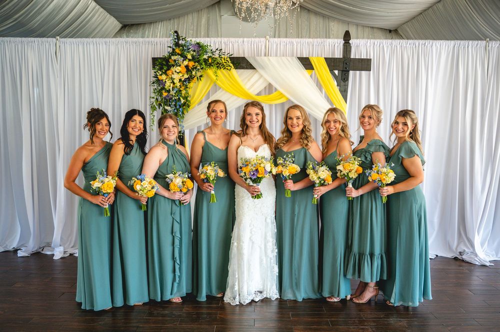 Bride in white lace gown with eight bridesmaids in teal dresses holding yellow and orange bouquets before floral backdrop.
