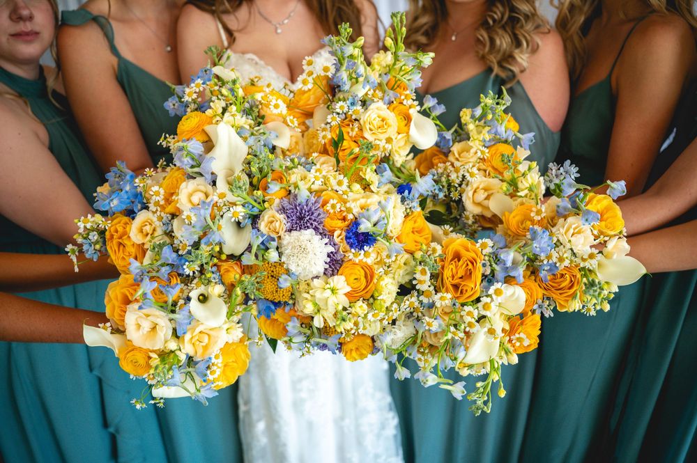 Bride and bridesmaids in teal dresses holding lush yellow, blue, and white floral wedding bouquets.