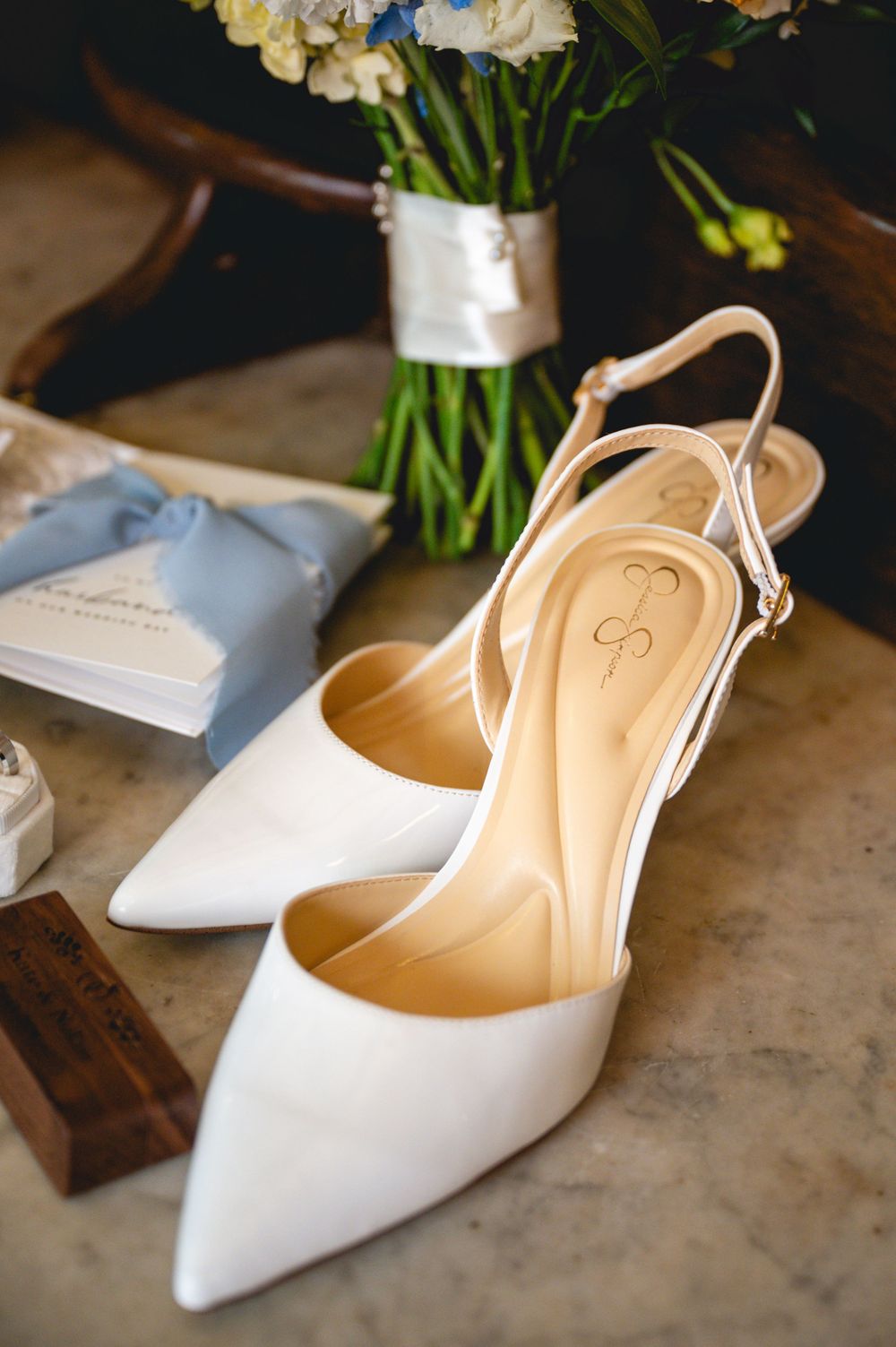 Elegant white pointed-toe slingback heels by Jessica Simpson displayed on marble surface beside bridal bouquet and accessories.