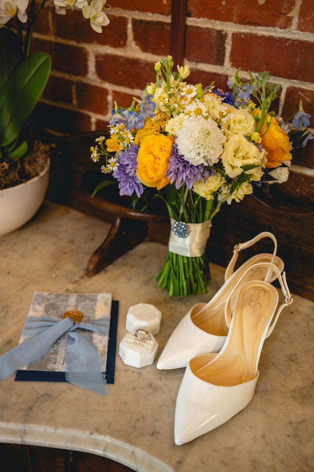 White bridal heels, wedding rings, blue gift box, and colorful bouquet with yellow, white, and purple flowers on marble table.