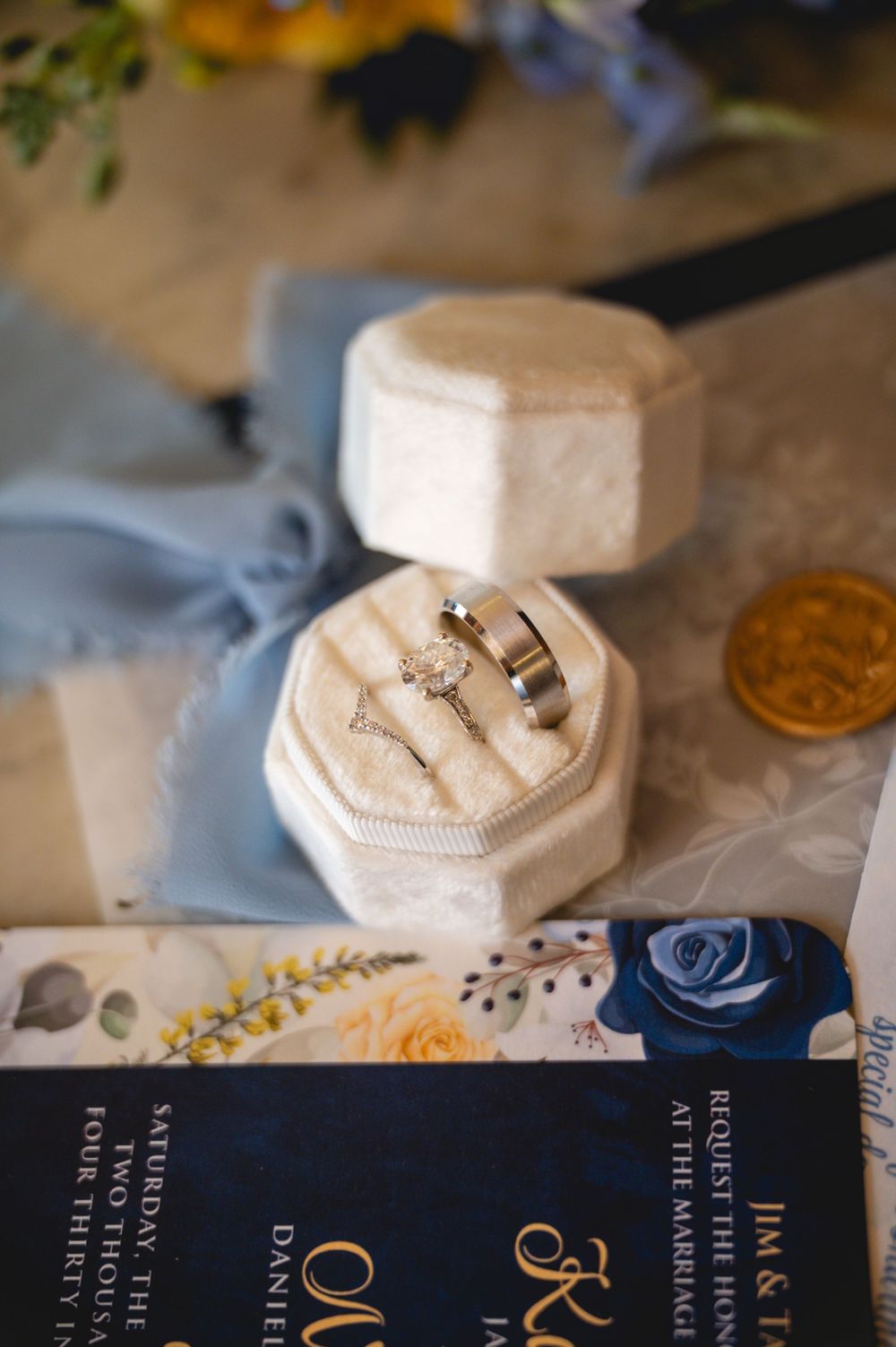 Elegant wedding rings displayed in an ivory velvet ring box atop navy blue floral wedding invitations and blue silk ribbon.