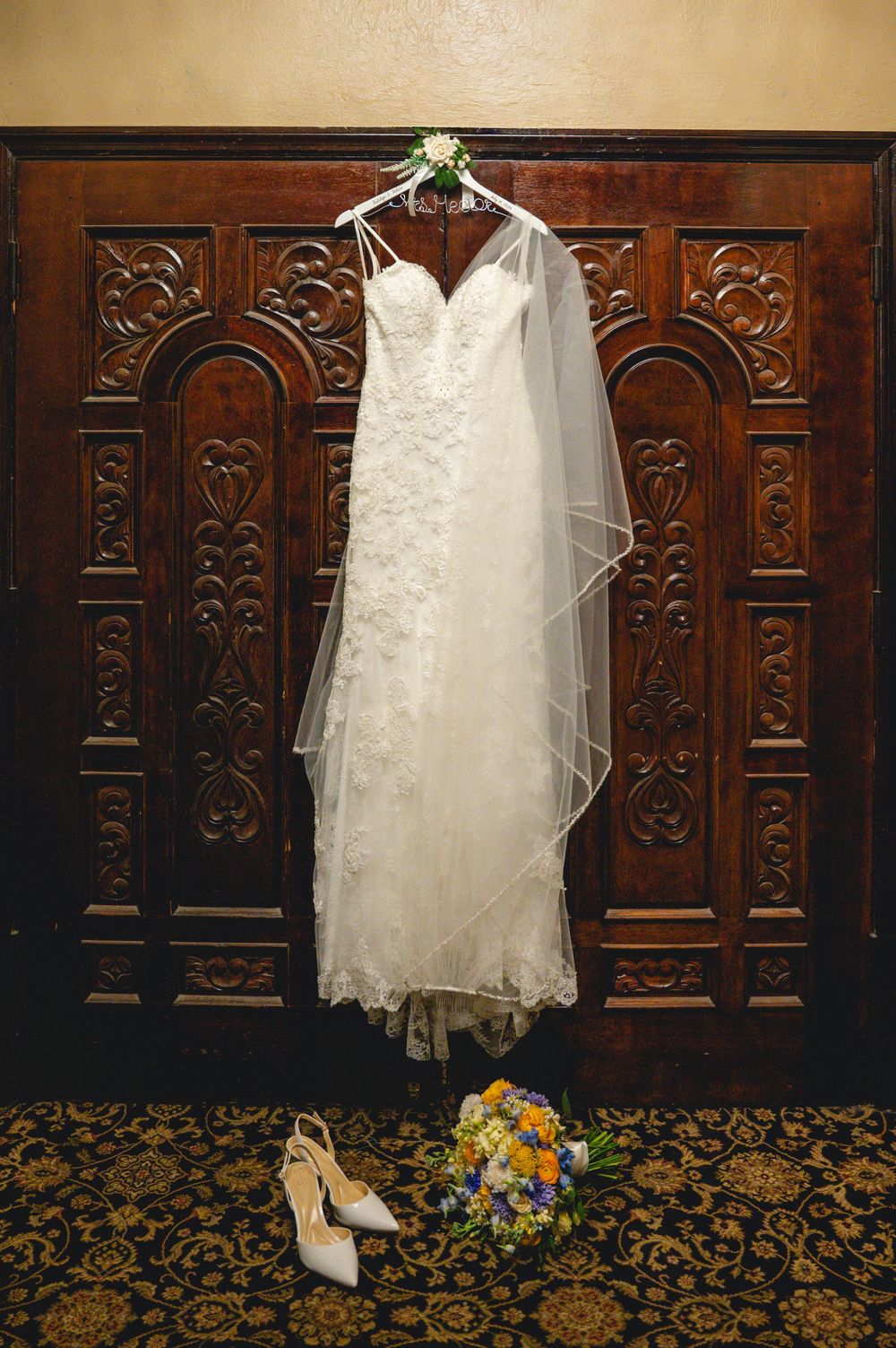 White lace wedding dress with veil hanging on ornate wooden doors, with bridal heels and colorful bouquet below.