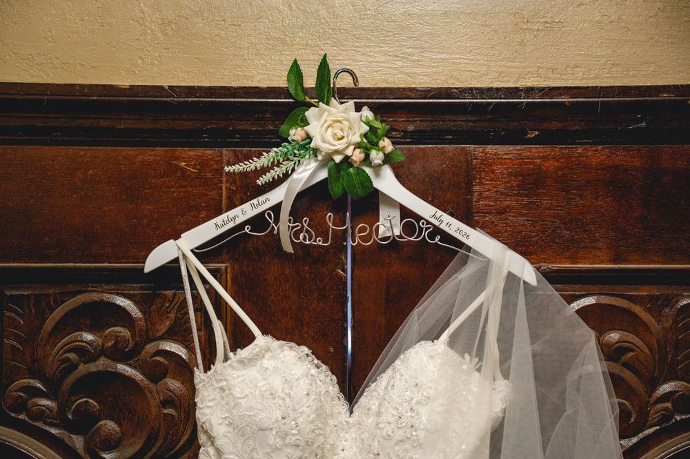 Wedding dress on a personalized Mrs. hanger with white roses and greenery, hung against a wooden wall with veil.