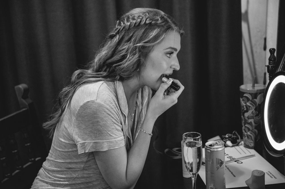 Black and white photo of a young woman with braided hair applying lipstick at a vanity mirror.