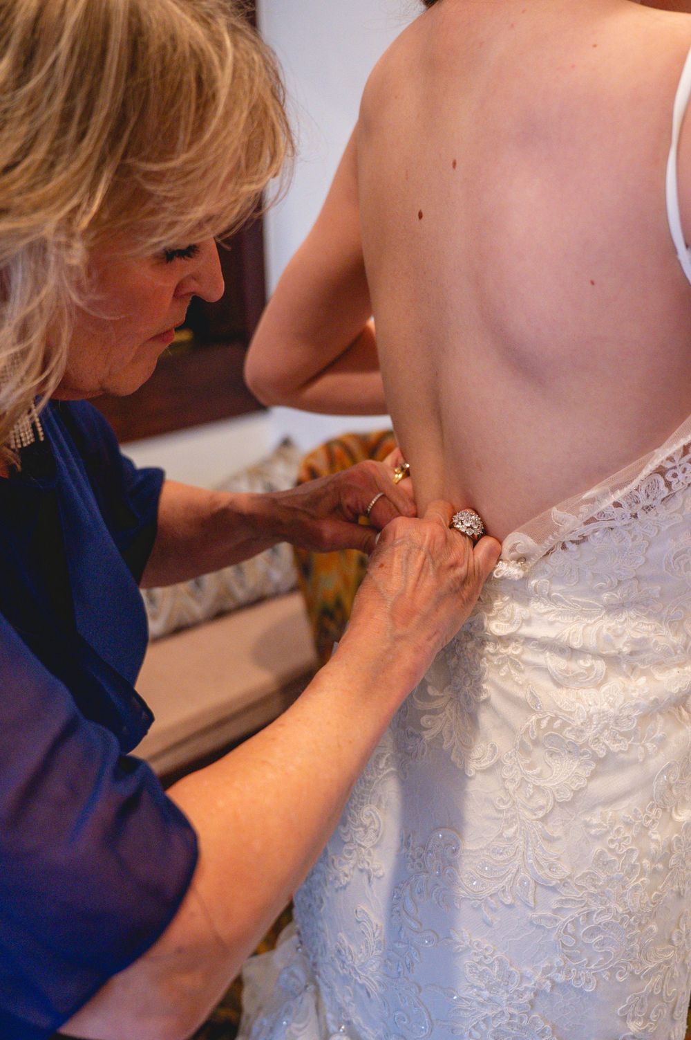 A woman in blue fastens the back buttons of a bride's white lace wedding dress before the ceremony.