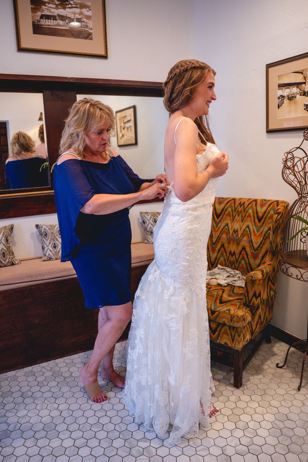 A bride in a white lace gown gets her dress fastened by a woman in a blue dress in an elegant bridal suite.