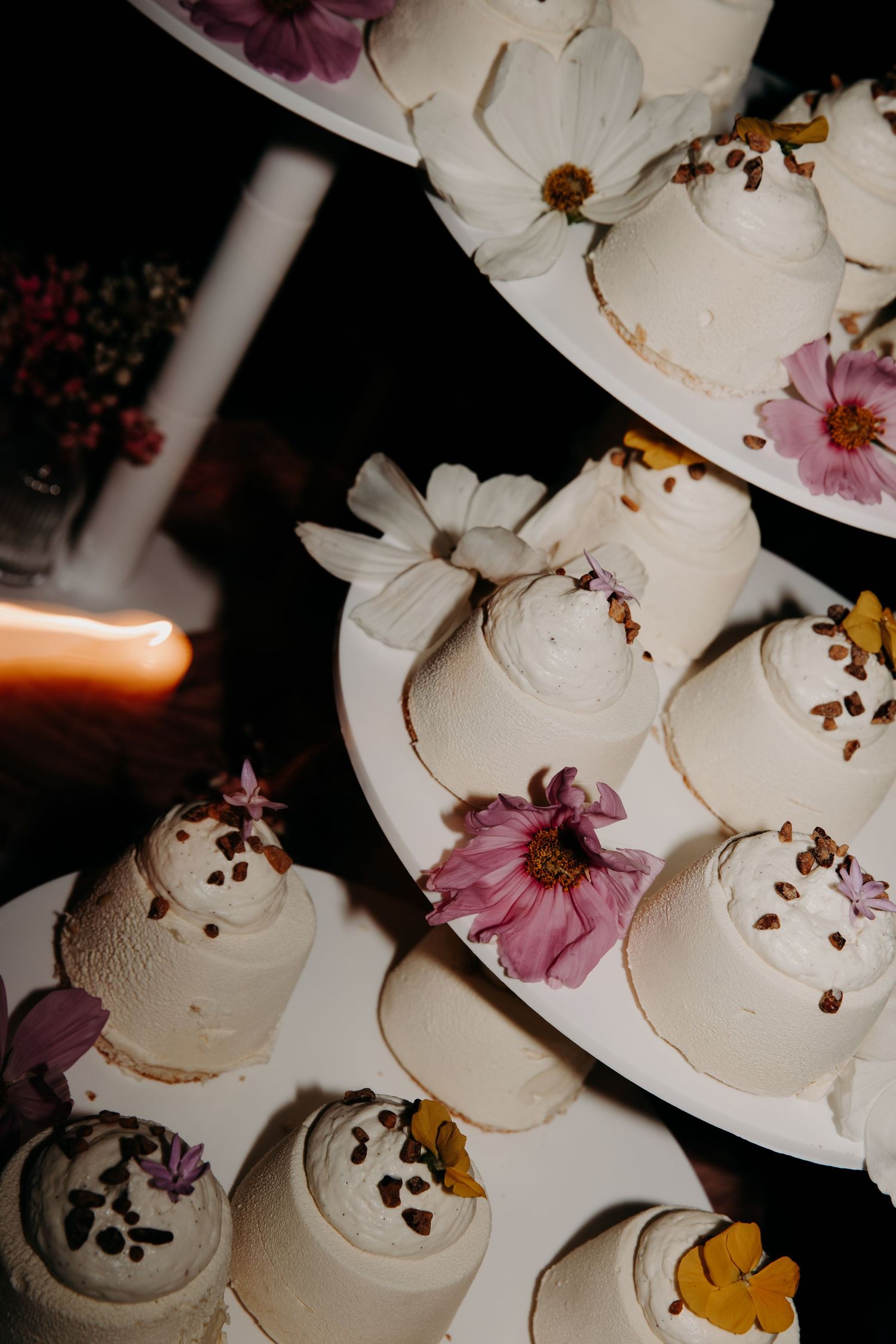 White frosted cupcakes decorated with fresh pink and yellow flowers displayed on a tiered stand at a wedding reception.