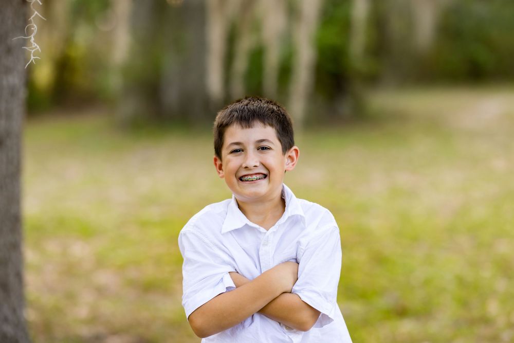 Smiling boy with braces in white shirt standing outdoors with arms crossed in a green park setting.