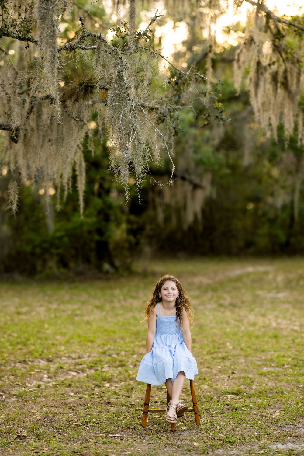 Young girl in a blue dress sitting on a wooden stool beneath Spanish moss-covered oak trees outdoors.