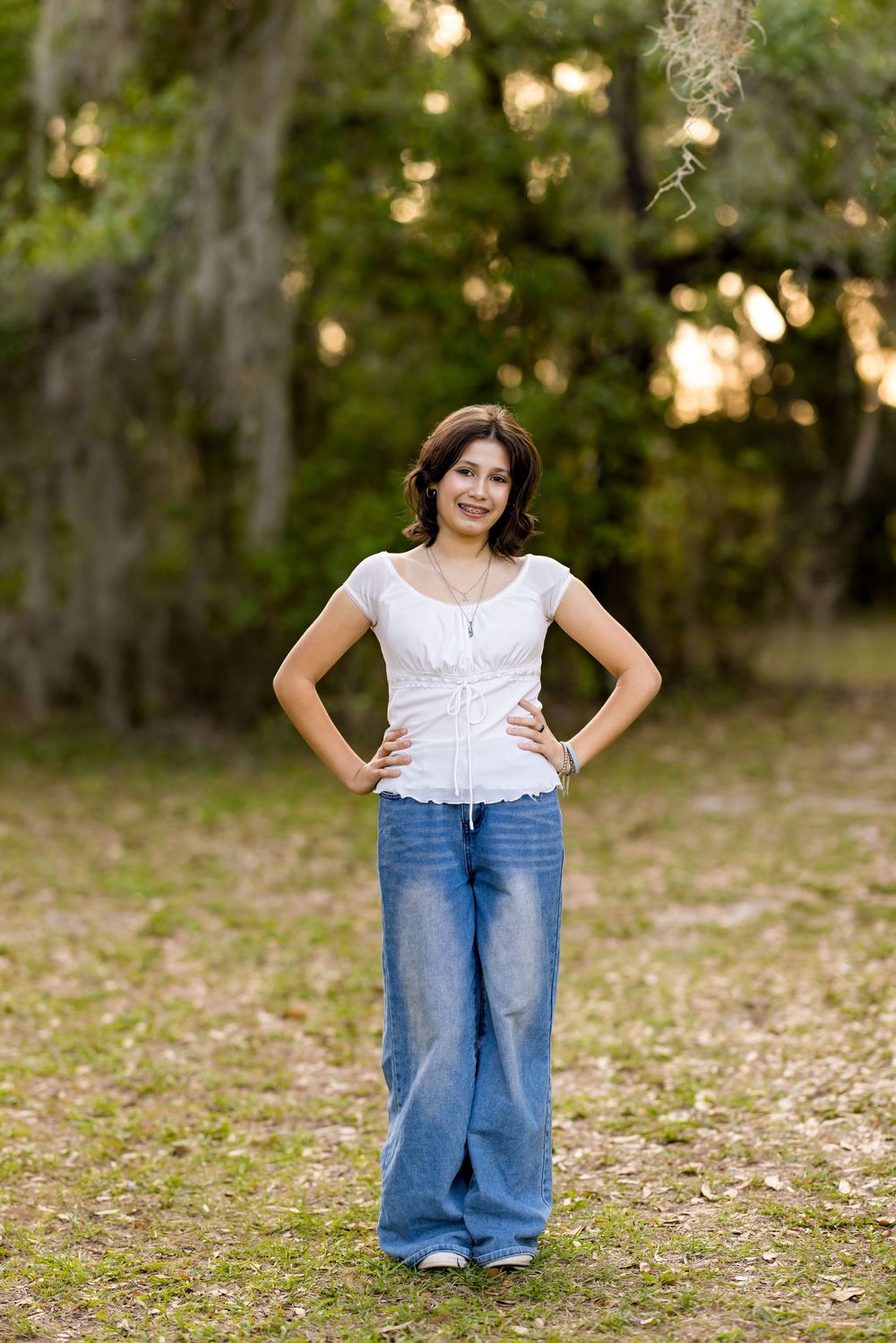 Teen girl in white top and wide-leg jeans standing confidently in a park with Spanish moss trees.