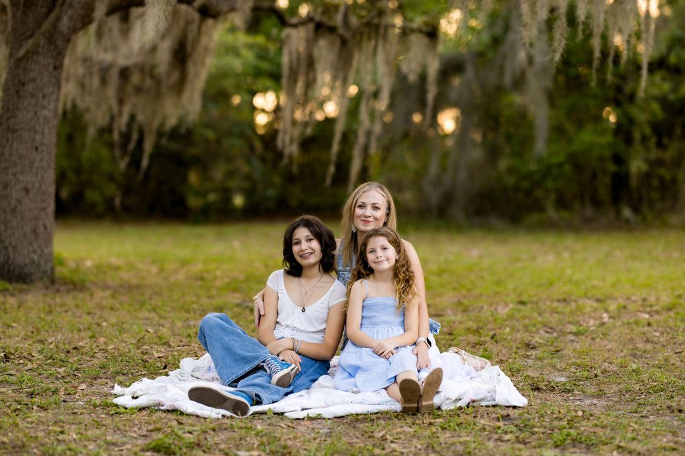Three females sitting on a white blanket under a moss-draped oak tree in a sunny park setting.