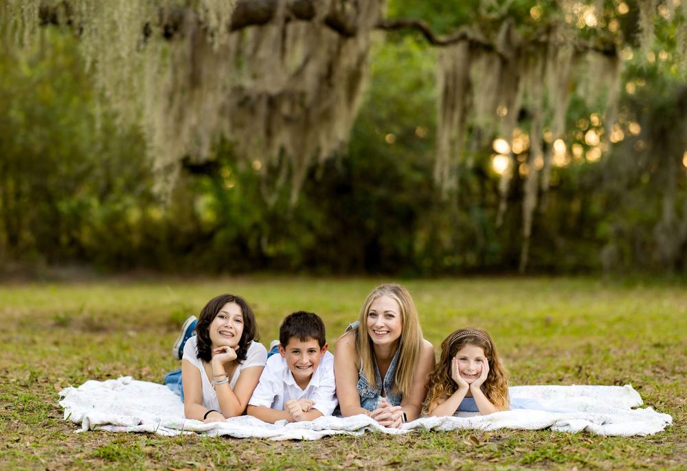Family of four lying on white blanket in grassy park with Spanish moss trees, smiling at camera.