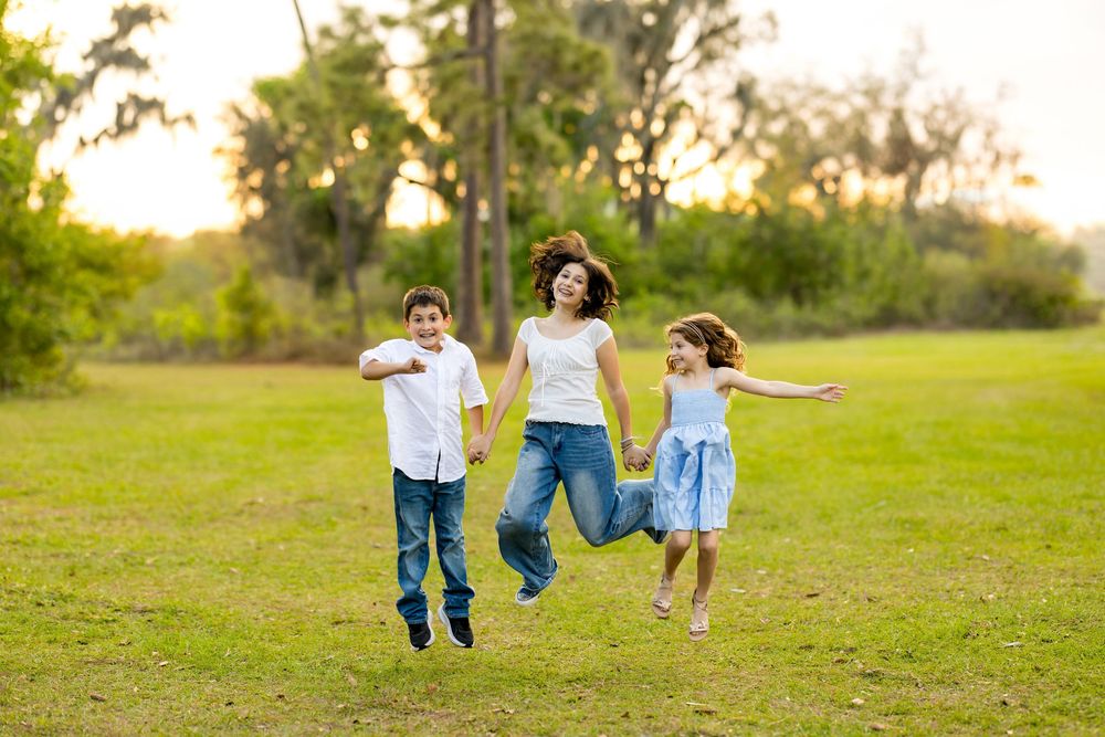 A joyful mother and two children jumping together in a lush green park at sunset.