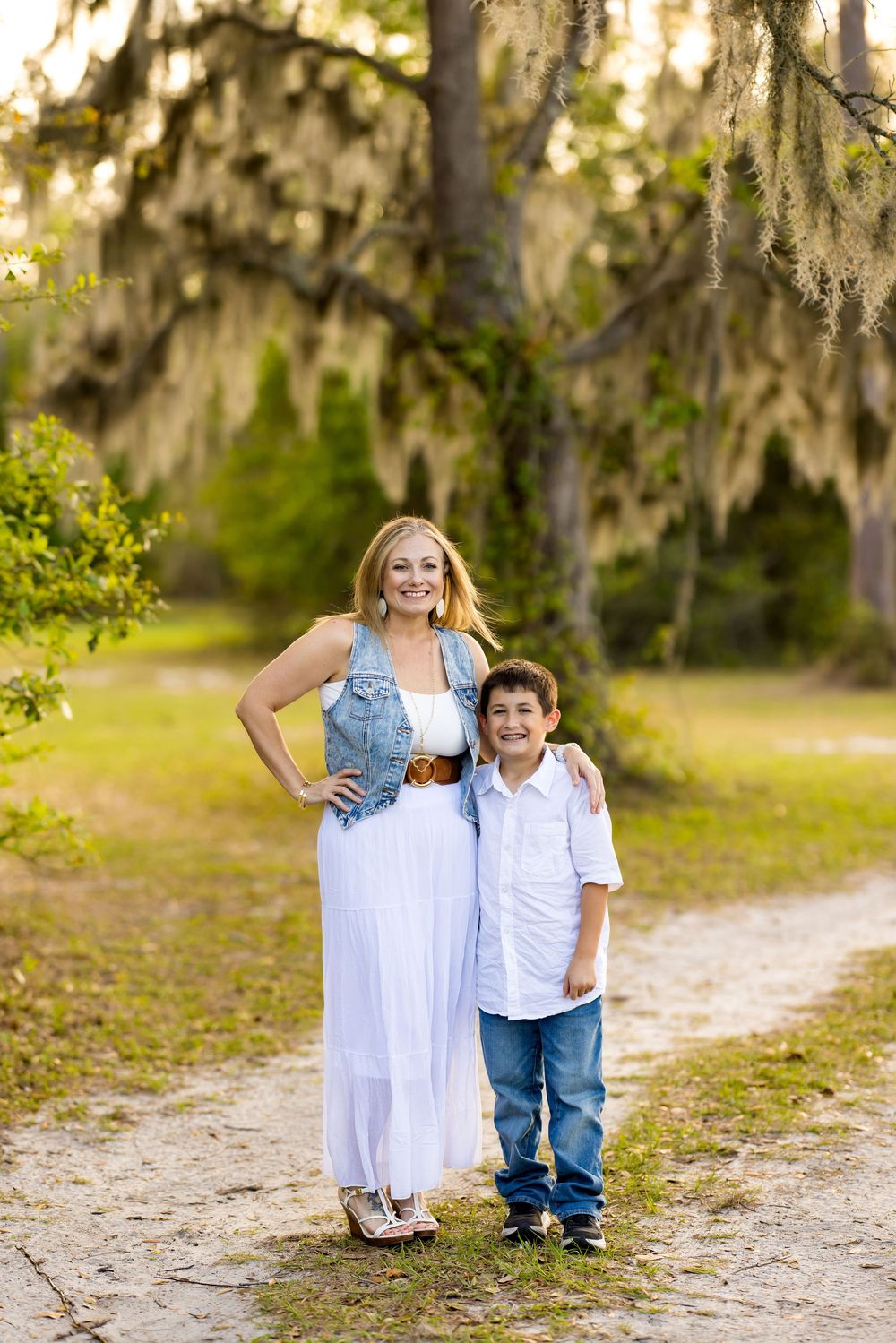 A woman in a white dress and denim vest poses with a young boy in jeans on a scenic outdoor path.