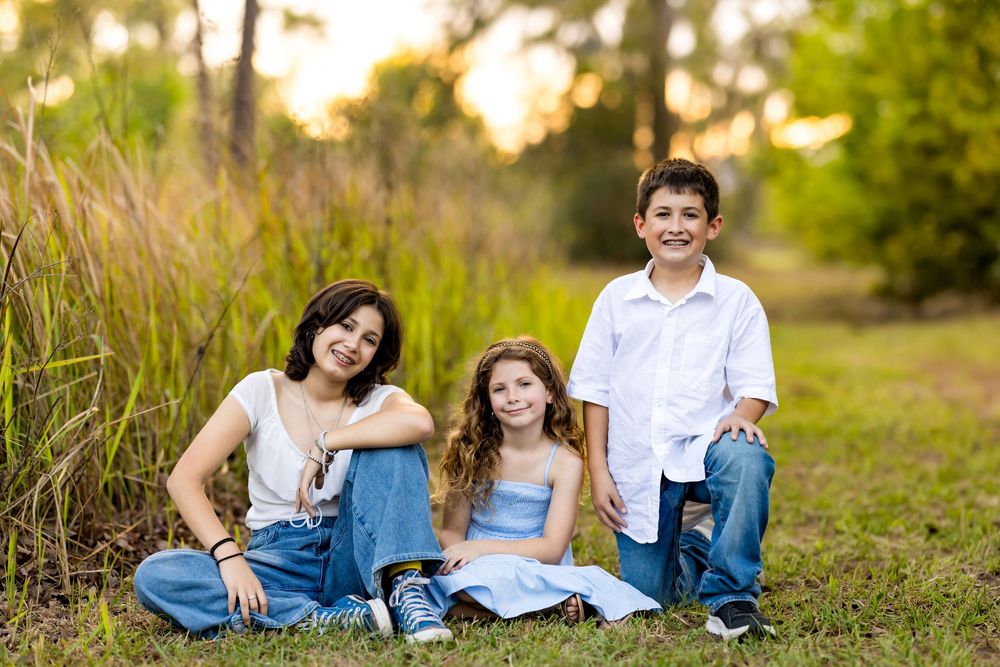 Three children smiling and posing outdoors in a grassy field during golden hour, wearing casual blue and white outfits.