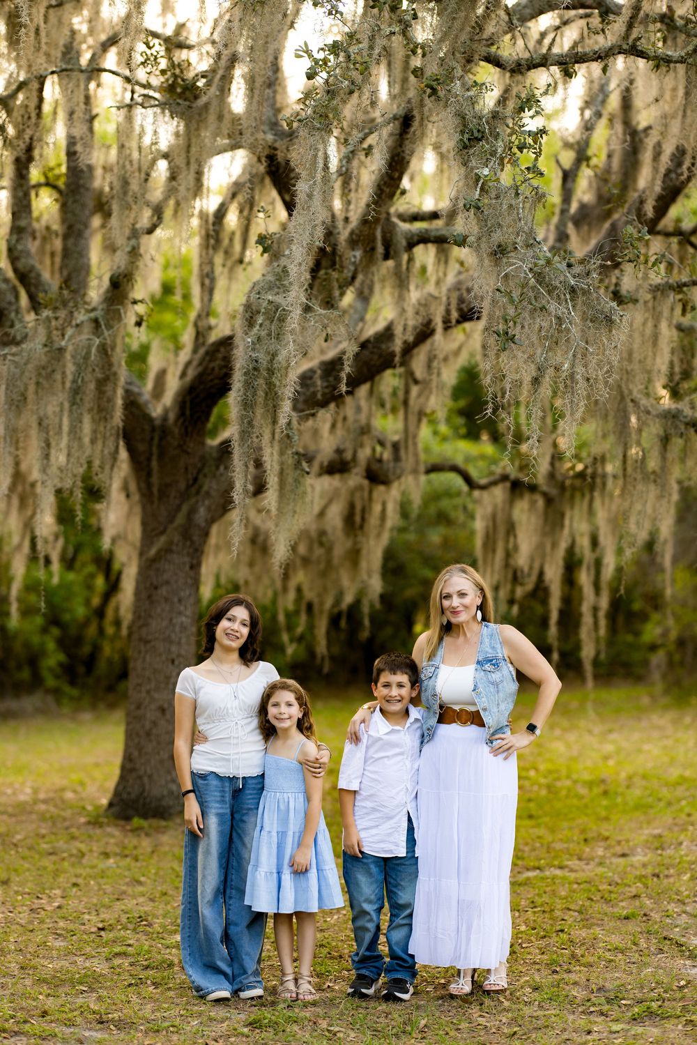 Family of four posing under a large Spanish moss-draped oak tree in a lush green park setting.