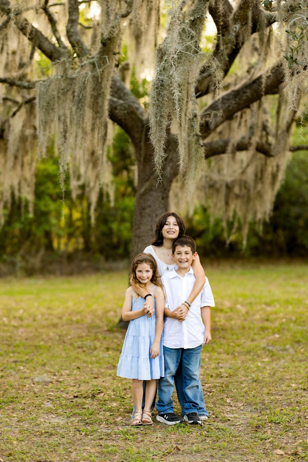 A mother and two children pose under a large Spanish moss-draped oak tree in a lush green park.