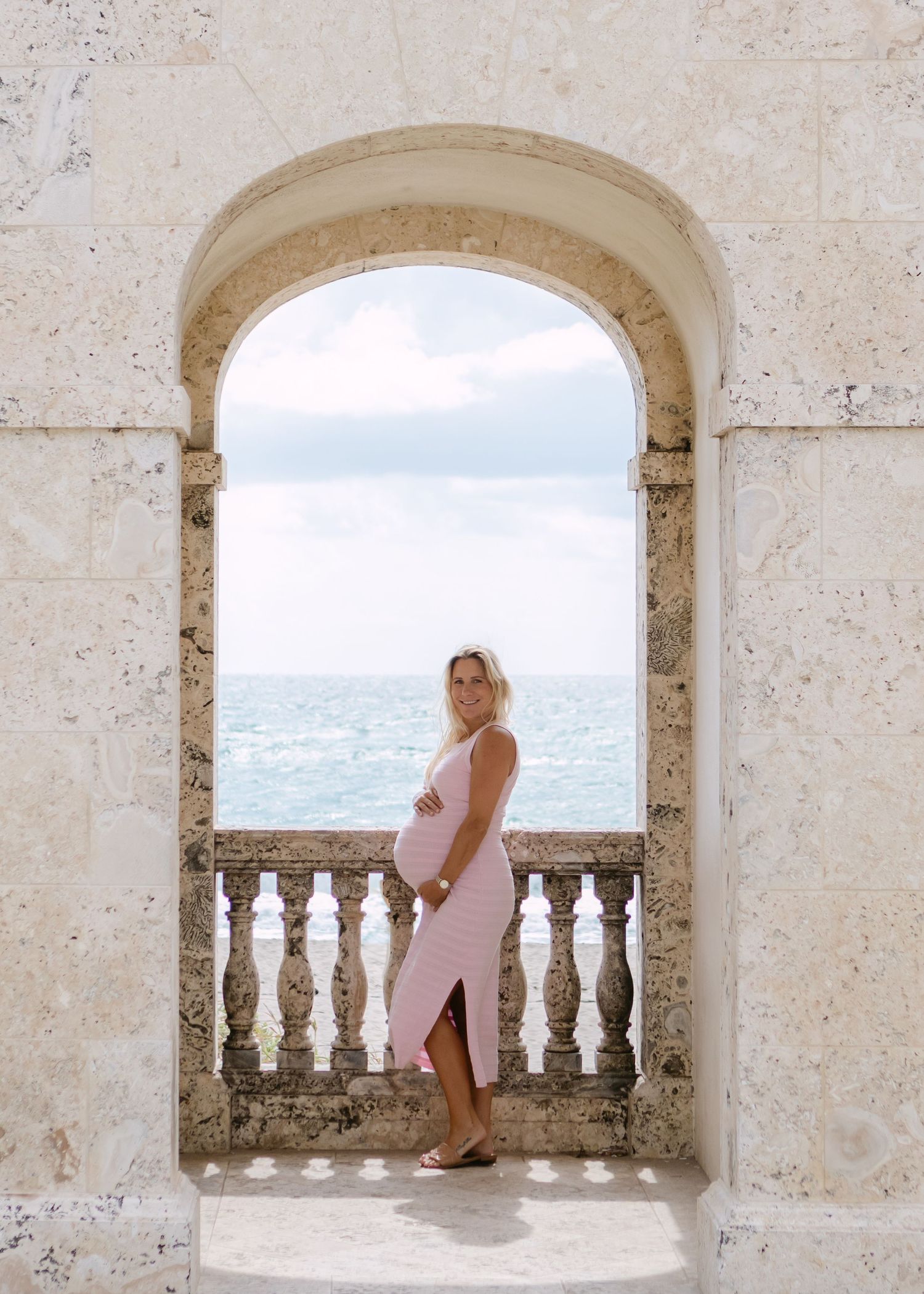 Pregnant woman in white lace off-shoulder dress cradling baby bump on beach at dusk.
