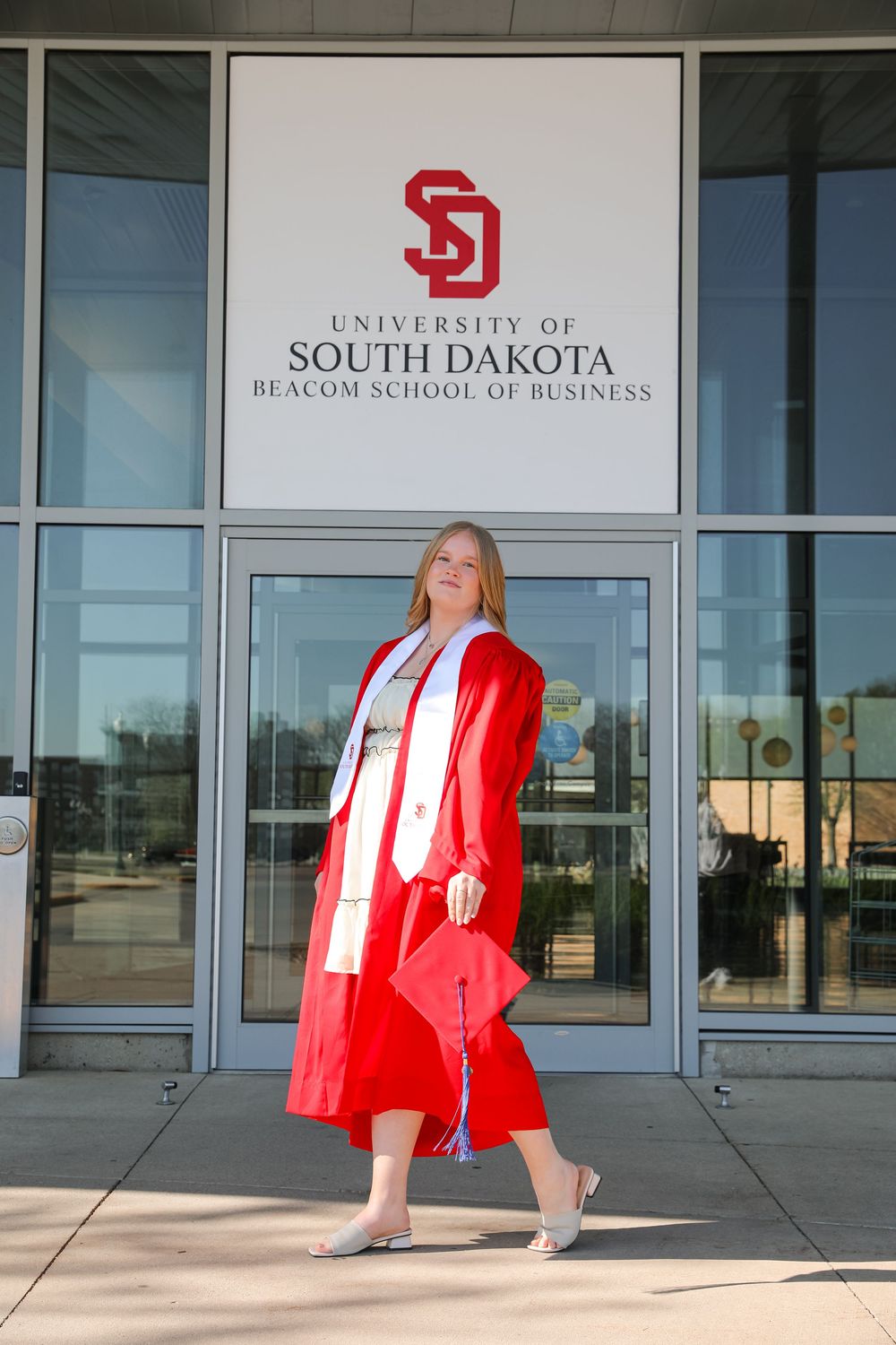 Graduate in red cap and gown poses outside University of South Dakota Beacom School of Business entrance.