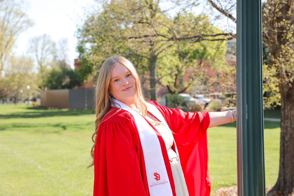 Young woman in red graduation gown and white stole posing outdoors on a sunny day on a green campus.