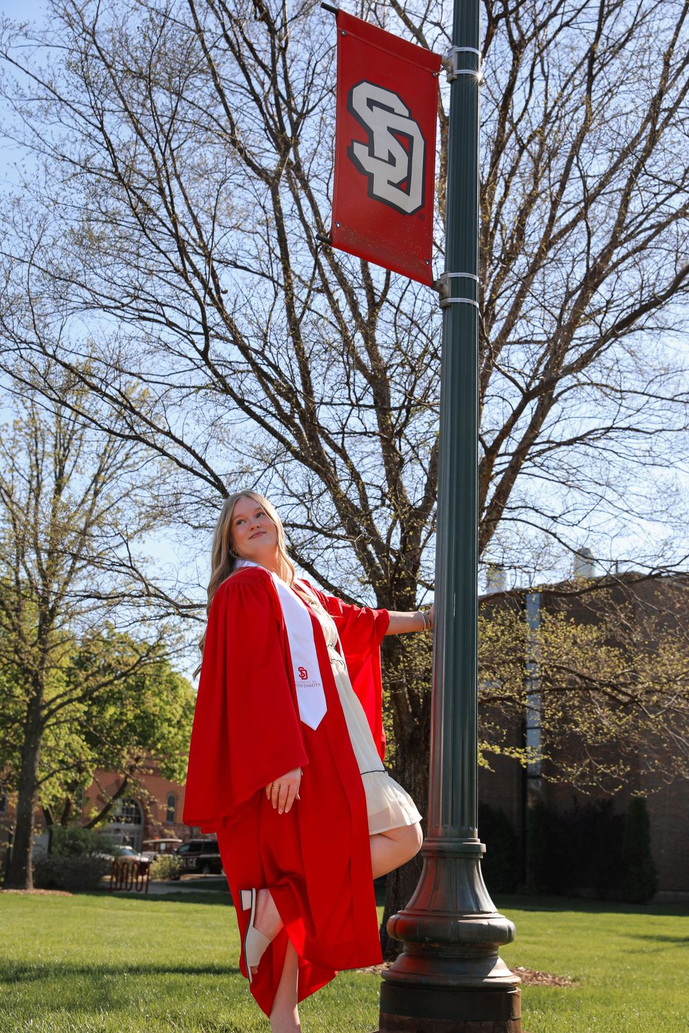 Blonde graduate in red cap and gown posing by University of South Dakota banner on campus lamppost.