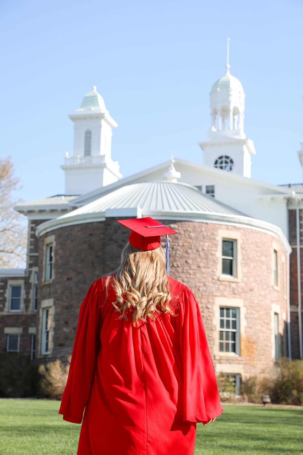 A blonde graduate in red cap and gown stands facing a historic stone building with white towers on a sunny day.